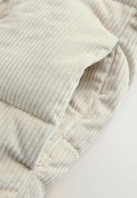 Helle beige Cordstoff mit vertikalen Streifen, präsentiert sich mit einer gesteppten Textur und weichem Finish, zeigt gefaltete Kanten und Nähte.