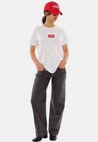 White t-shirt with red logo, gray wide-leg pants, and a red cap with white text. The outfit features clean lines and casual styling.