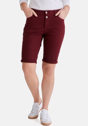 Person trägt hochgekrempelte, burgunderrote knielange Shorts, ein weißes Langarmshirt und weiße Turnschuhe, mit den Händen in den Taschen.