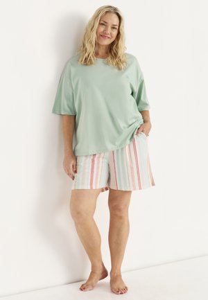 Kvinde i mintgrøn oversized t-shirt og pastelfarvede stribede shorts står barfodet, smilende, lænet mod en hvid væg med den ene hånd i lommen.