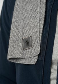 Grå uldhalstørklæde med et struktureret hørfiskemønster; har en sort rektangulær lap med logo. Bæres over en marineblå zip-up jakke.
