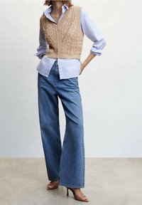 Beige gestrickte Weste mit V-Ausschnitt über einem hellblauen Hemd, kombiniert mit weiten blauen Jeans und hellbraunen Absätzen.