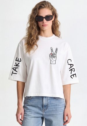 Femme portant des lunettes de soleil noires, t-shirt blanc avec un signe de paix fait avec la main et les mots "Faith," "Love," "Peace," "Joy," "Hope," et "Take Care" sur les manches.