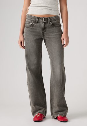 Personne portant un jean large gris délavé, un top court blanc et des chaussures plates rouges, debout sur un fond clair.