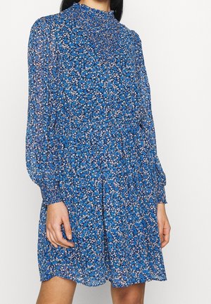 Femme portant une robe bleue fleurie à manches longues avec un col smocké haut et des poignets, debout devant un fond uni.