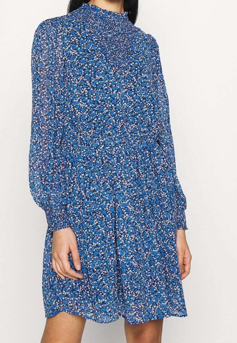 Femme portant une robe bleue fleurie à manches longues avec un col smocké haut et des poignets, debout devant un fond uni.