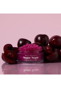 Rosa burk med läppskrubb och texturerad magentafärgad produkt inuti, omgiven av mörkröda körsbär på en ljusrosa bakgrund.
