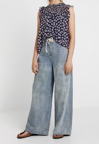 Top senza maniche floreale blu navy con spalline arricciate, abbinato a jeans a gamba larga blu chiaro. Scarpe casual completano l'outfit.