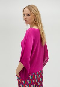Haut léger de couleur rose vif avec des manches courtes et larges. Présente une coupe décontractée et un col simple, associé à un pantalon coloré à motifs.