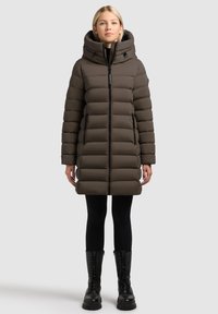 Brązowa pikowana parka z wysokim kołnierzem i kapturem, z przednim zamkiem błyskawicznym i dwoma bocznymi kieszeniami, w zestawieniu z czarnymi legginsami i butami.