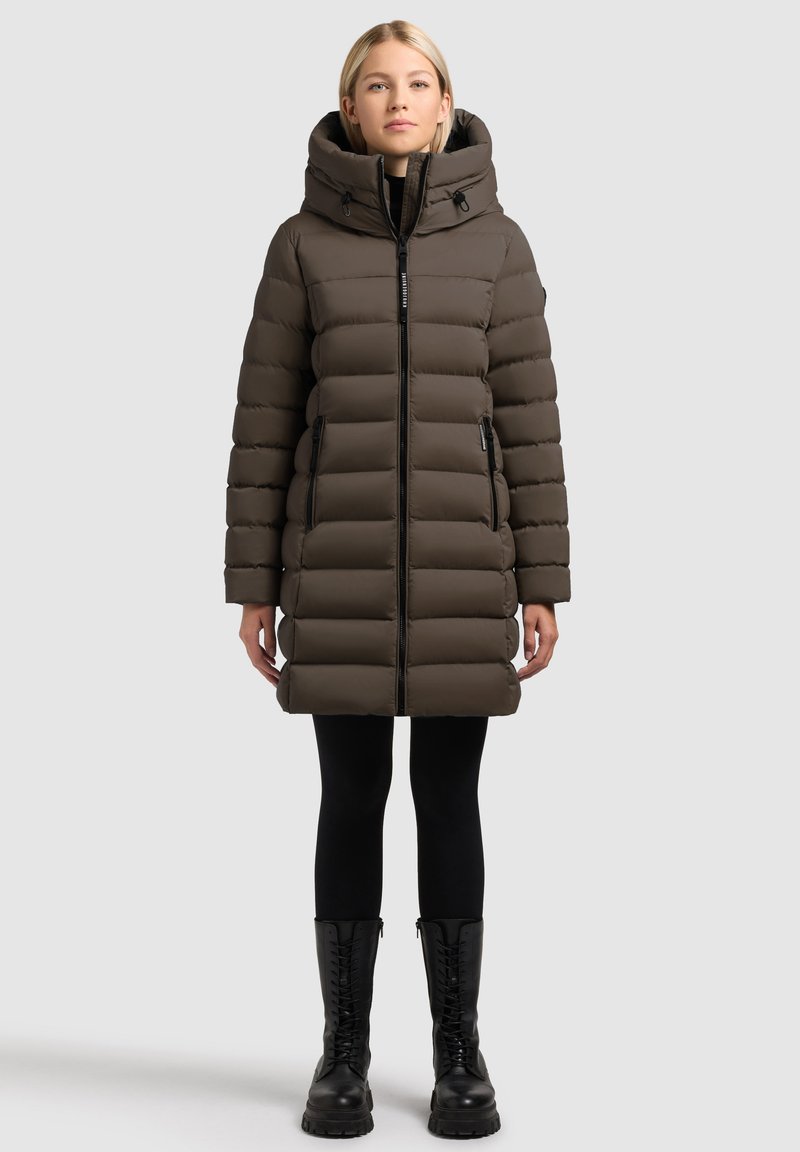 Brązowa pikowana parka z wysokim kołnierzem i kapturem, z przednim zamkiem błyskawicznym i dwoma bocznymi kieszeniami, w zestawieniu z czarnymi legginsami i butami.