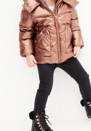 Enfant portant une doudoune brune brillante, des leggings noirs et des bottes d'hiver noires, debout devant un fond blanc.