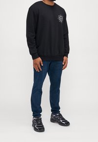 Sudadera negra con un gráfico de sol en el lado izquierdo del pecho, combinada con vaqueros azules y zapatillas negras con detalles morados.