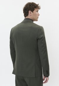 Alyvučių žalumos pritaikyta švarko struktūra su lygiu paviršiumi, griežtu siluetu ir subtiliomis siūlėmis, su juodais mygtukais ir iškarpytu apykakle.