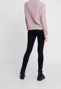 Personne debout de dos, portant un pull en maille rose clair, un jean skinny noir et des bottes noires à boucle, sur un fond uni.