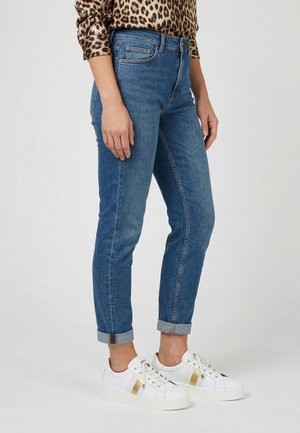 Kvinde iført opkrogede blå jeans, hvide sneakers med guldstriber og en leopardprint bluse med lange ærmer, stående mod en ensfarvet baggrund.