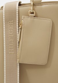 Borsa beige con una superficie liscia, caratterizzata da una cerniera dorata e un portacarte removibile. La tracolla presenta dettagli intrecciati con accenti di un beige chiaro.