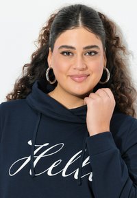 Felpa con cappuccio blu navy con testo bianco in corsivo che dice "Ciao". Presenta lacci e polsini a coste; il modello indossa grandi orecchini a cerchio.