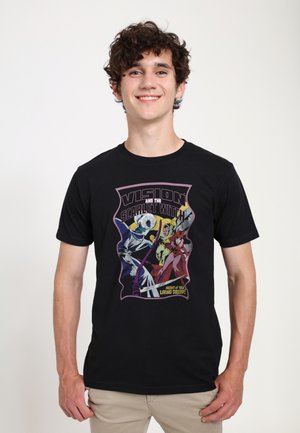 Junger Mann lächelt, trägt ein schwarzes T-Shirt mit farbenfrohem Comic-Stil-Motiv "Vision and the Scarlet Witch" und beige Hosen.