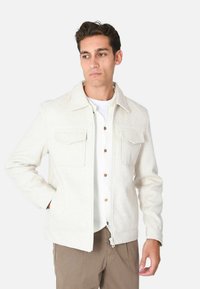 Giacca a zip color crema realizzata in tessuto testurizzato. Presenta due tasche sul petto e un colletto classico. Abbinata a una camicia bianca e pantaloni marroni.