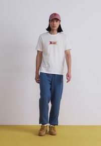 T-shirt blanc avec un patch logo coloré, pantalon de survêtement bleu et chaussures beiges. Le modèle porte une casquette rose ; fond minimaliste.