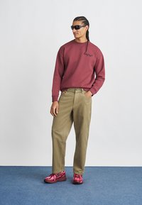 Sweat-shirt marron avec logo sur le devant, pantalon kaki et baskets roses brillantes. Modèle portant des lunettes de soleil noires, se tenant devant un fond clair.