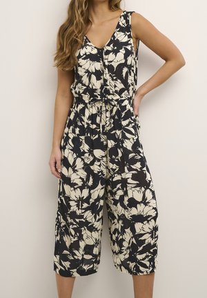 Kobieta ubrana w bezrękawnikowy kombinezon w czarno-biały kwiatowy wzór z dekoltem w serek i szerokimi, krótkimi nogawkami, pozuje z jedną ręką na biodrze.