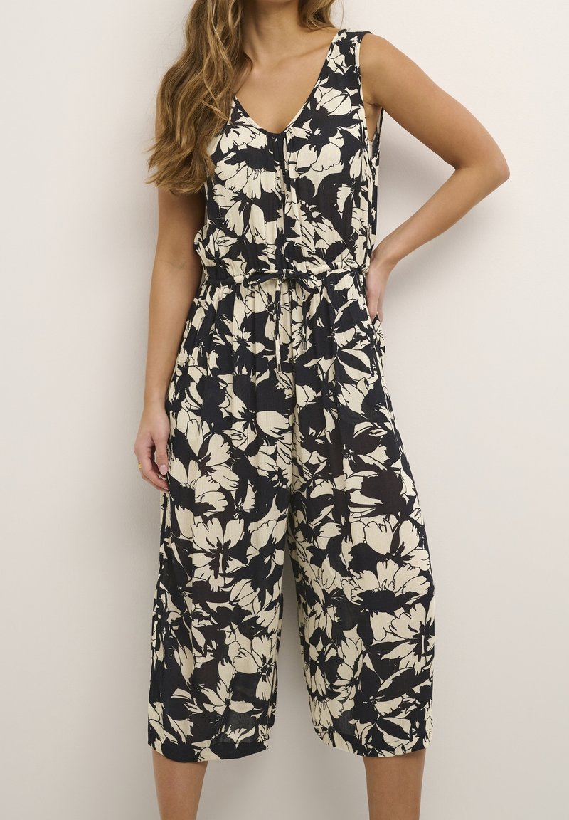 Femme portant une combinaison sans manches noire et blanche à motif floral, avec un décolleté en V et un pantalon large raccourci, posant avec une main sur la hanche.