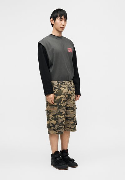 Γκρι και μαύρο πουκάμισο με μακριά μανίκια με το λογότυπο "ISOD"· κάπρι παντελόνι καμουφλάζ με τσέπες· μαύρα ψηλά αθλητικά παπούτσια με λουριά.