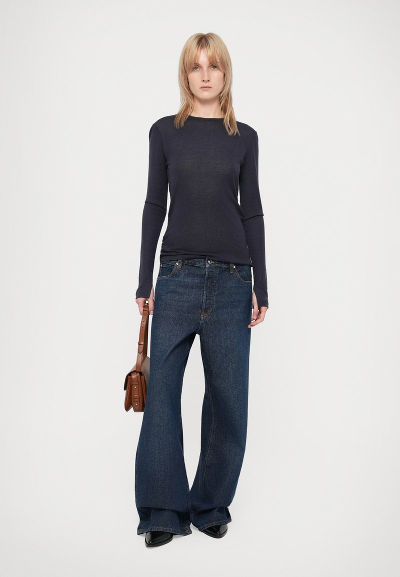 Marineblaue Langarmbluse kombiniert mit hellen, weit geschnittenen Jeans. Das Model hält eine braune Umhängetasche. Die Textur wirkt glatt, der Denim hat minimale Details.