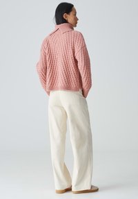 Rosa gestrickter Pullover mit erhabener Textur, hohem Kragen und lockerer Passform, kombiniert mit cremefarbenen, weit geschnittenen Cordhosen. Einfache Schuhe sind zu sehen.
