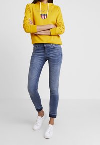 Henkilö, joka käyttää keltaista hupparia logolla, sinisiä kapeita farkkuja, joissa on repaleiset lahkeet, ja valkoisia lenkkareita, seisoo kädet ristissä.