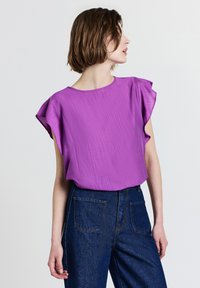 Top corto viola con finitura texturizzata e spalline arricciate, abbinato a jeans in denim blu a vita alta con tasche posteriori.