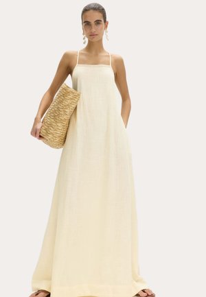 Kvinde i en cremefarvet maxikjole med spaghetti-stropper, der holder en stor vævet stråclutch og bærer hængende øreringe.
