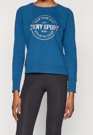 Persoon draagt een blauw DKNY Sport sweatshirt met de tekst "New York City Athletic Club 1989" en zwarte legging met zijlogo detail.