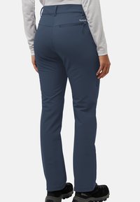 Pantalons de randonnée bleu marine en tissu extensible avec une coupe slim. Équipés d'une poche arrière zippée et de poignets renforcés, associés à des chaussures noires.