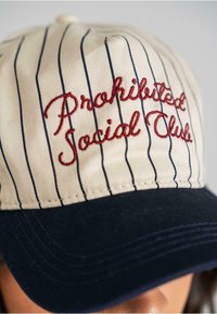 Baseballcap i crème og marineblå farver med lodrette marineblå striber og rød broderet tekst, der står "Prohibited Social Club."