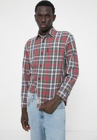 Chemise à manches longues à carreaux dans des tons rouges, bleus et gris, avec un col boutonné et une poche poitrine unique, associée à un jean bleu clair.