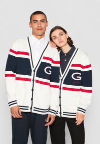 Hvit strikket cardigan med marineblå, rød og sølvfargede striper. Har et stort "G"-logo, knappelukking og frontlommer.