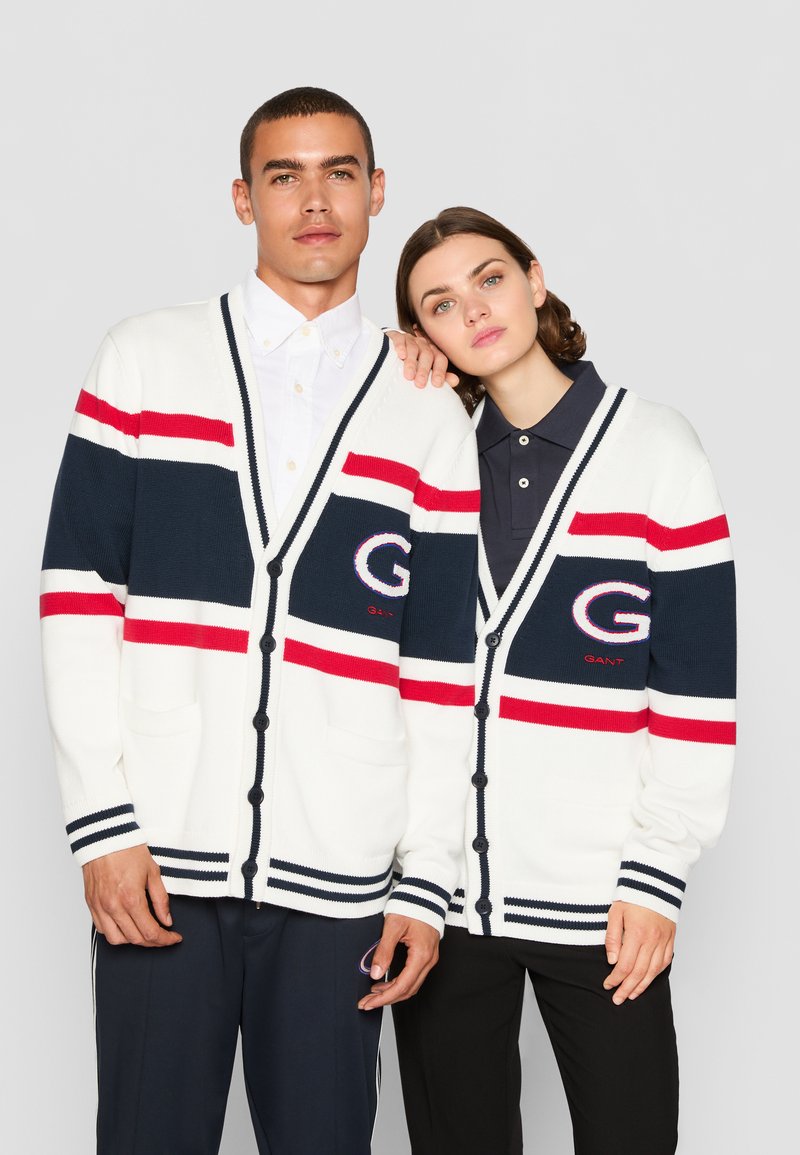 Hvit strikket cardigan med marineblå, rød og sølvfargede striper. Har et stort "G"-logo, knappelukking og frontlommer.