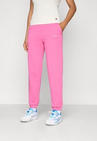 Pink sweatpants with elastic cuffs, side pockets, and embroidered logo detail. Worn with white and blue athletic shoes.