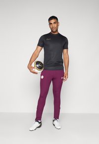 Melnā īsā piedurkņu sporta krekls ar zelta logotipu, apvienots ar ribotām purpura biksēm ar logotipu, un melnbaltu futbola bumbu.
