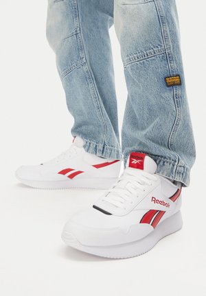 Białe sneakersy Reebok z czerwonymi akcentami i logo, z teksturowaną cholewką i płaską podeszwą, w zestawieniu z jasnoniebieskimi dżinsami.