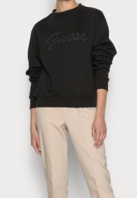 Czarna bluza z bawełny o mieszanym składzie, z okrągłym dekoltem i długimi rękawami. Posiada wyszywane logo "Guess" z przodu. W zestawie z beżowymi spodniami.