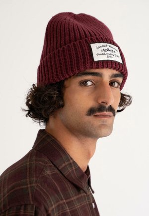 Man with curly hair and mustache wears a maroon knit beanie and matching plaid shirt, looking sideways against a plain background.