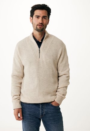 Beige strikket sweater med halv-zip krave og ribbet struktur, parret med blå jeans. Stoffet ser blødt og hyggeligt ud i en afslappet pasform.
