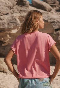 Top de manga corta rosa con bordes ondulados, hecho de una tela suave, combinado con shorts de colores claros y estampados, con un fondo rocoso.
