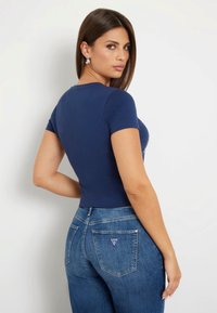 Tamsiai mėlynas trumpomis rankovėmis bodysuit'as su prigludusia forma, derinamas su vidutinio aukščio mėlynomis džinsomis, su trikampio logotipu ant užpakalinės kišenės.