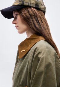 Νεαρή γυναίκα με μακριά καστανά μαλλιά φοράει πράσινο μπουφάν με καφέ γιακά από κοτλέ και μια καρό καπέλο με σκοτεινό γείσο, κοιτάζοντας προς τα αριστερά.