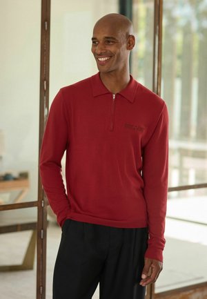 Smiling bald man wearing a red long-sleeve zip-up shirt and black pants, standing indoors near glass doors with his hand in his pocket.